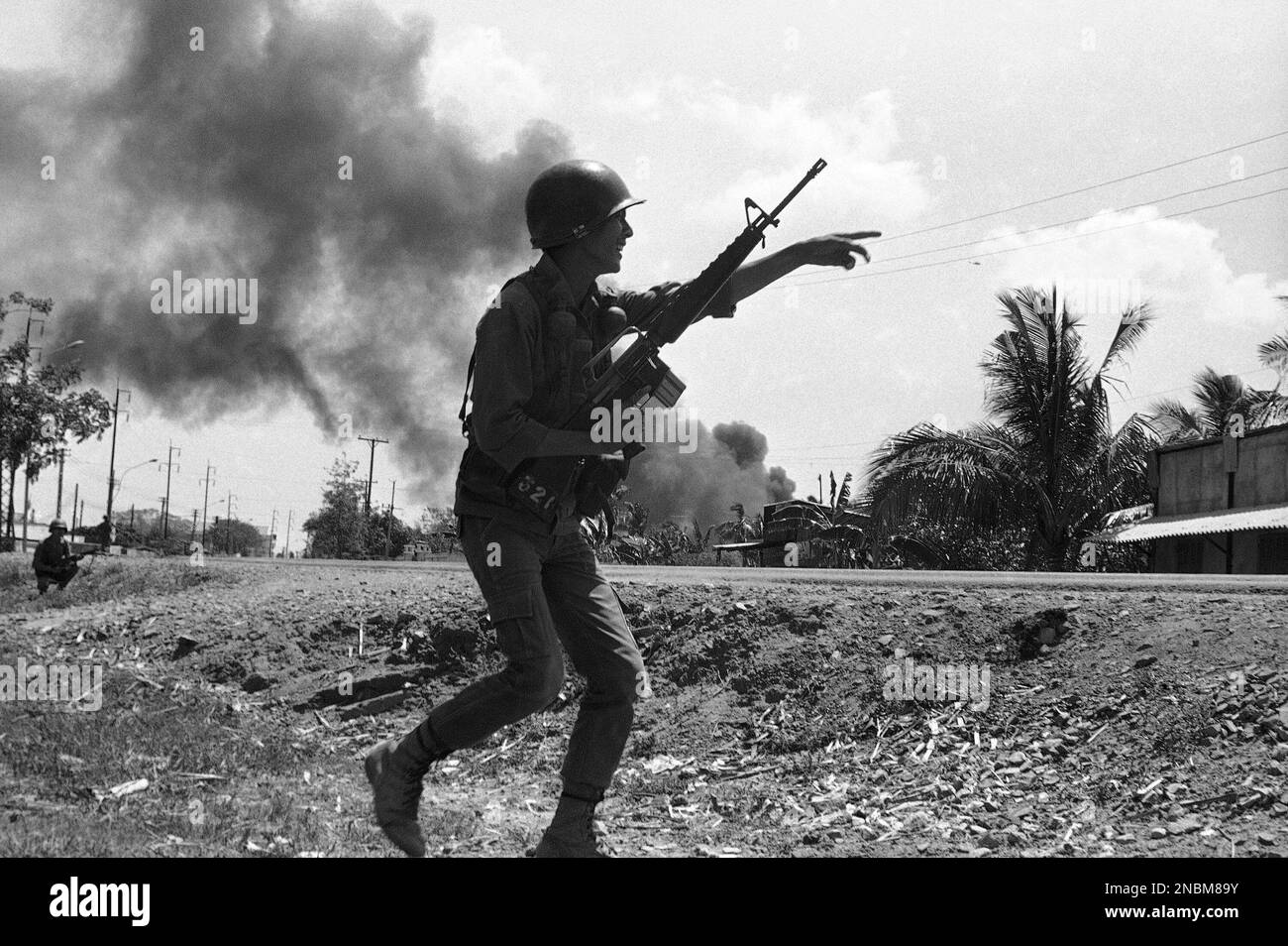 A South Vietnamese soldier gestures to a buddy as he runs along Route 4 in Tan An, South Vietnam on April 10, 1975, during a battle. Black smoke behind him comes from gas storage tank hit by Communist shells. (AP Photo/Lien) Stock Photo