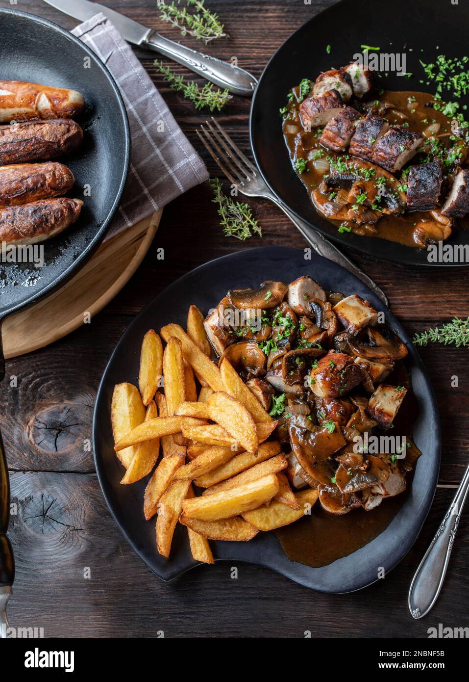 Fried sausage dish with pan fried german bratwurst, onion mushroom sauce and homemade french fries on a table with plates and pan. Flat lay Stock Photo