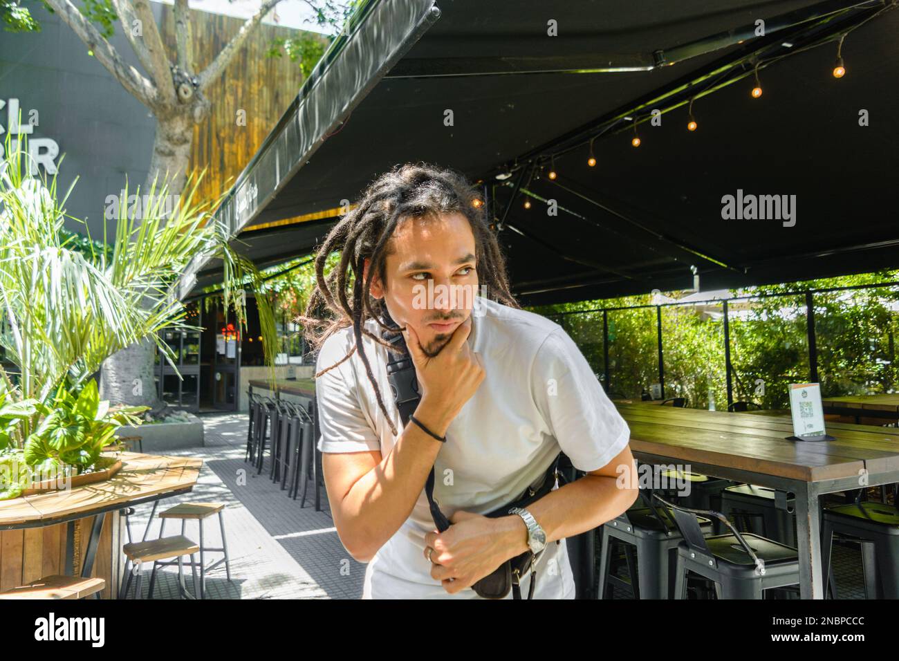 young latin venezuelan man with dreadlocks, standing outside a restaurant touching his chin and looking doubtfully to the side Stock Photo