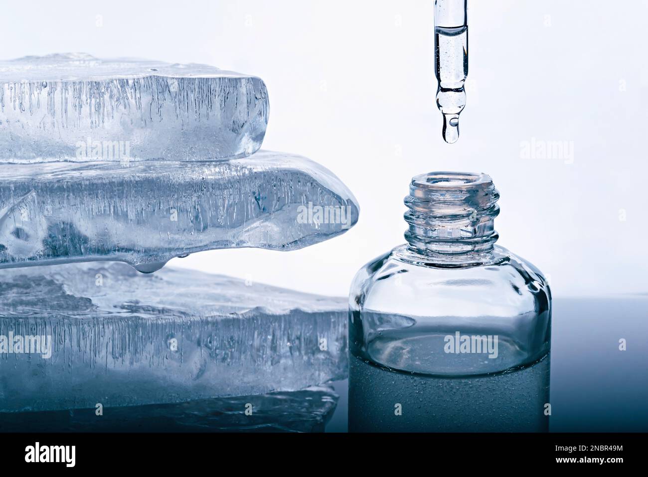 A bottle of face serum and a pipette with a falling drop on the white background with pieces of melting ice. Stock Photo