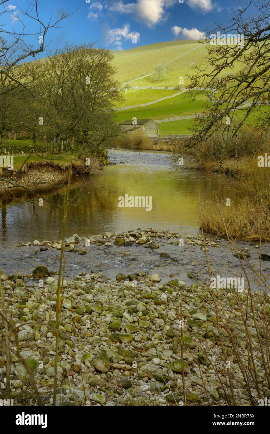 On a beautiful sunny day in Kettlewell, North Yorkshire,  sunlit rolling hills reflect on The River Wharfe with pebbles and rocks in the shallow water. Stock Photo