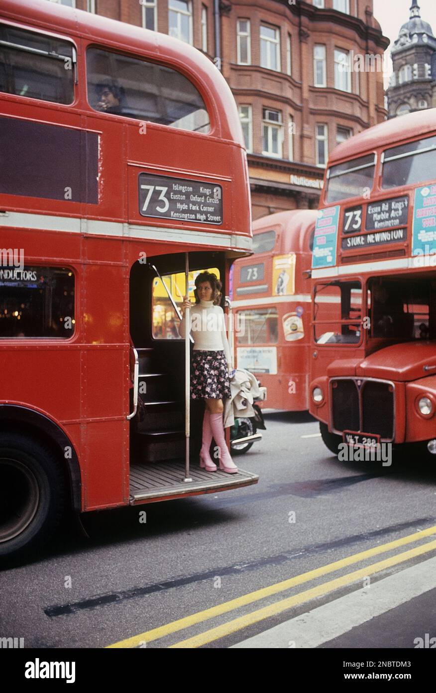 London 1972. A street view of Oxford street with the typical red double decked buses. A young woman is seen on the platform ready to go off. She is dressed in a typical 1970s outfit, jumper, short pink patterned skirt and matching pink leather boots in a platform model. A shoe fashion that at this time was popular in Europe and Britain and continued to be so until 1976 when platform shoes suddenly went out of fashion. This year 1972 famous people like David Bowie, David Johansen of the New York Dolls wore platform shoes.  Kristoffersson ref DV2 Stock Photo