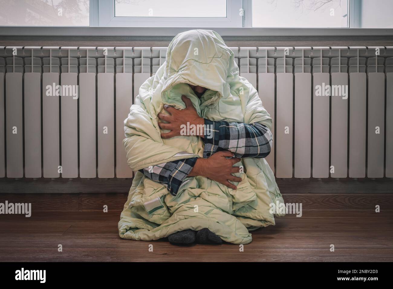 It's cold at home in wintertime. Man freezing in his house in winter because of broken thermostat. Sad young guy wrapped in plaid shivering while sitt Stock Photo