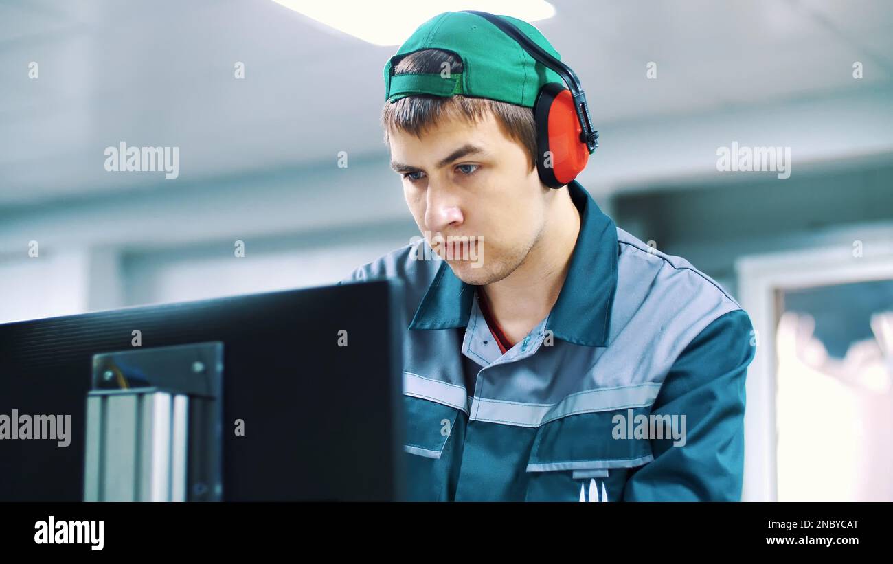 portrait of an employee of an enterprise, factory. a worker in red headphones from noise, looks at a computer monitor, adjusts, controls work of machines, automated production. High quality photo Stock Photo