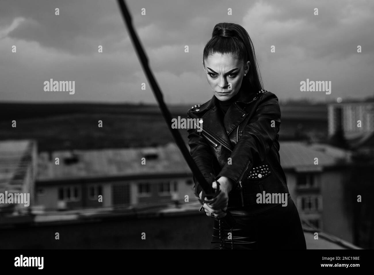 A grayscale of female in black ninja assassin costume holding a sword and standing near wall Stock Photo