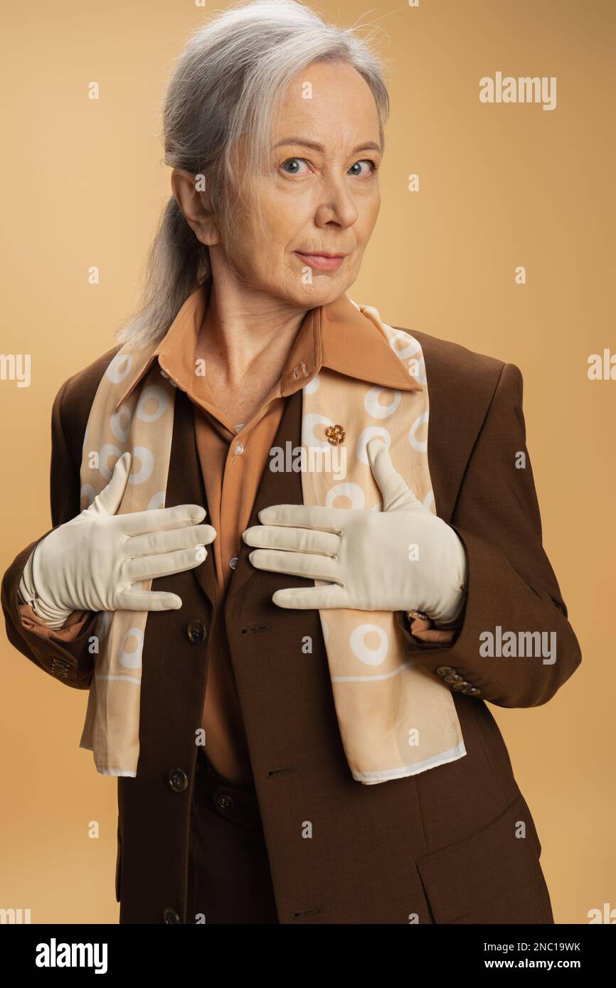 senior woman in brown formal wear and white gloves posing isolated on beige Stock Photo