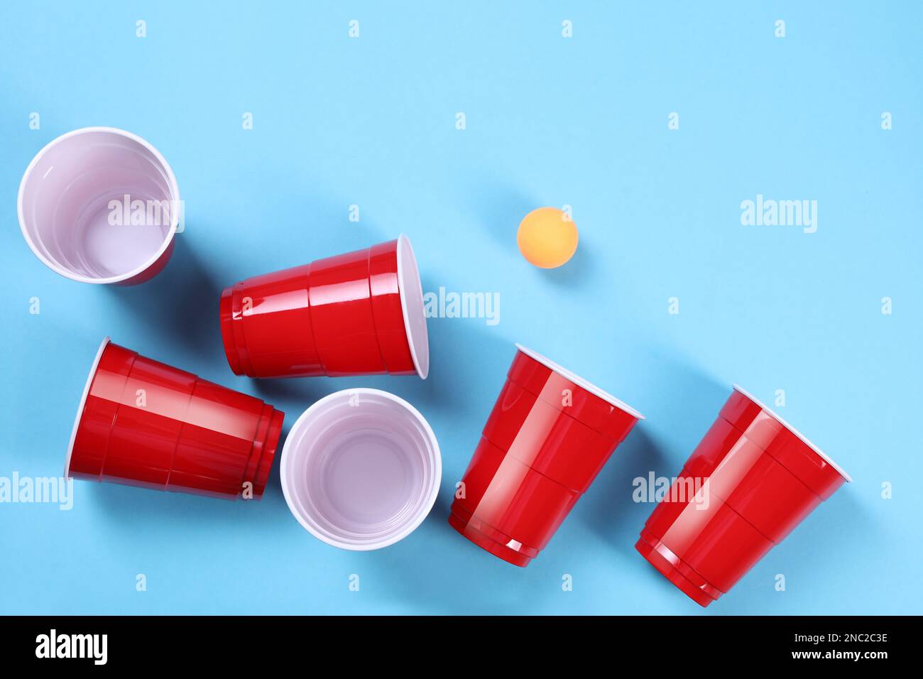 Plastic cups and ball on light blue background, flat lay. Beer pong