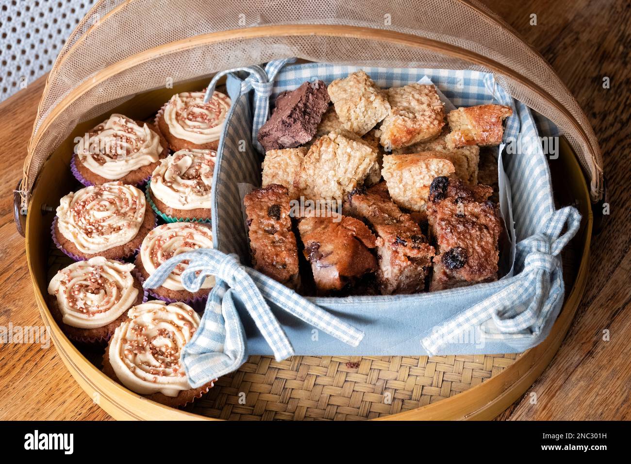 An assortment of tasty cake treats including cup cakes, Flapjacks and slices of Bread Pudding. All are displayed in a netted bamboo food cover Stock Photo