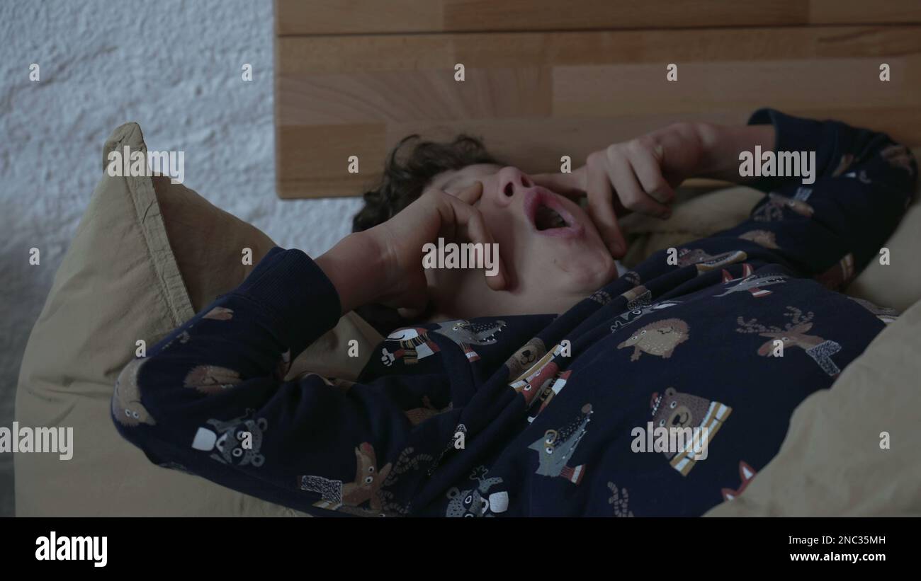 One young boy laying in bed yawning in the morning. Kid waking up rubbing face and eyes