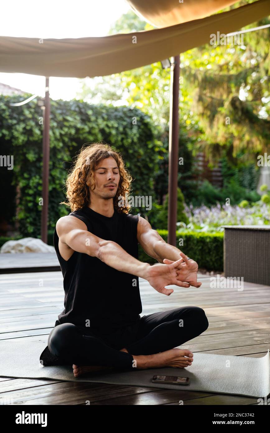 Young curly man doing workout while sitting on exercise mat in yoga studio outdoors Stock Photo