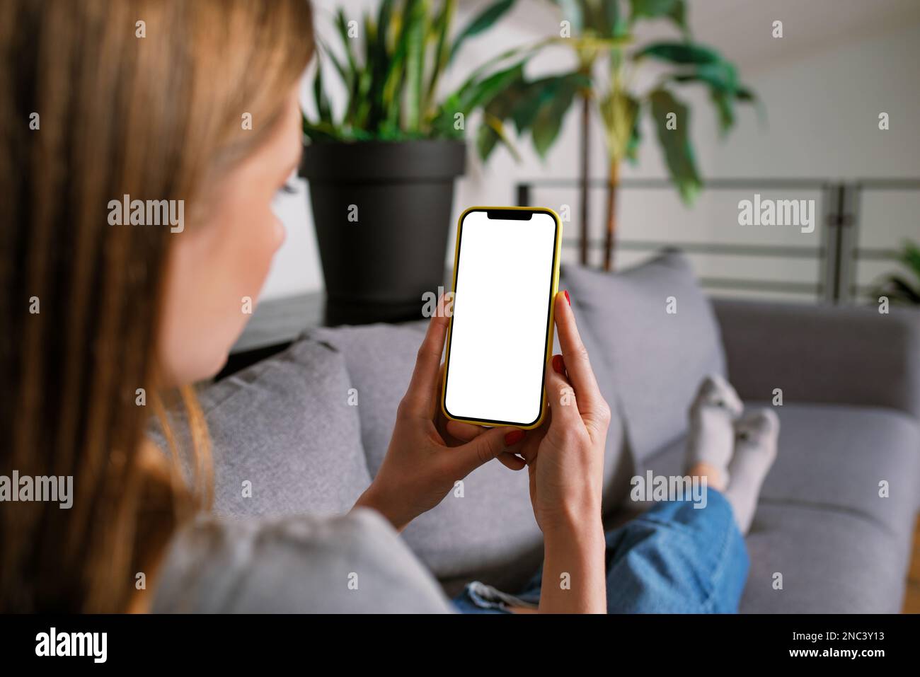Back view an Unrecognizable Casual blonde young woman in a modern living room lying a sofa working with iPhone smartphone mockup with a blank screen Stock Photo