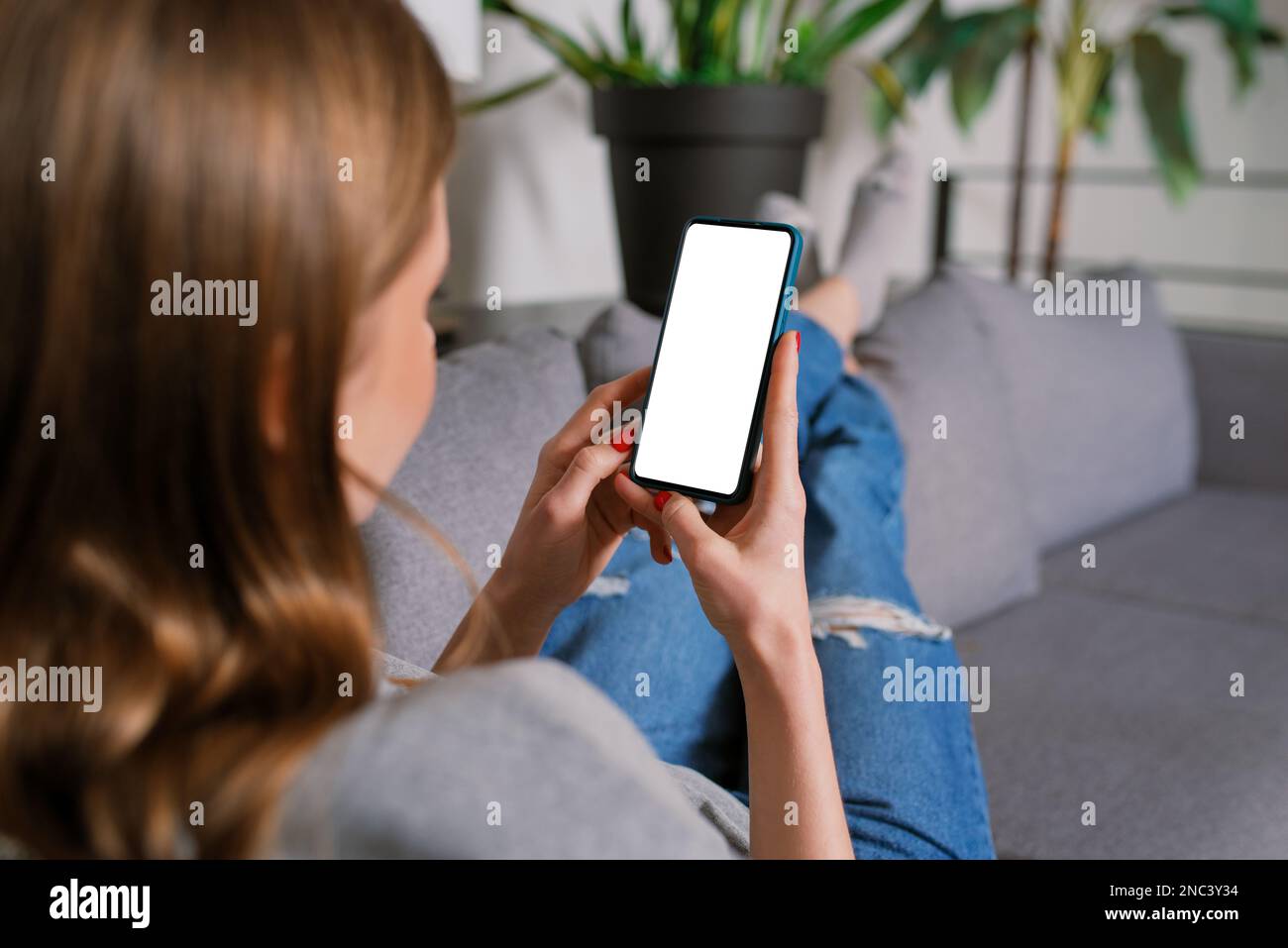 Back view an Unrecognizable Casual blonde young woman in a modern living room lying a sofa working with iPhone smartphone mockup with a blank screen Stock Photo
