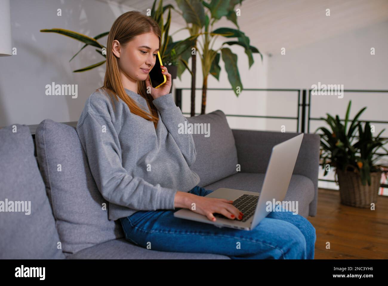 Cute young woman talking on her smartphone a looking a modern laptop screen sitting on the cozy sofa at home, Hands at work with Digital Technology Stock Photo