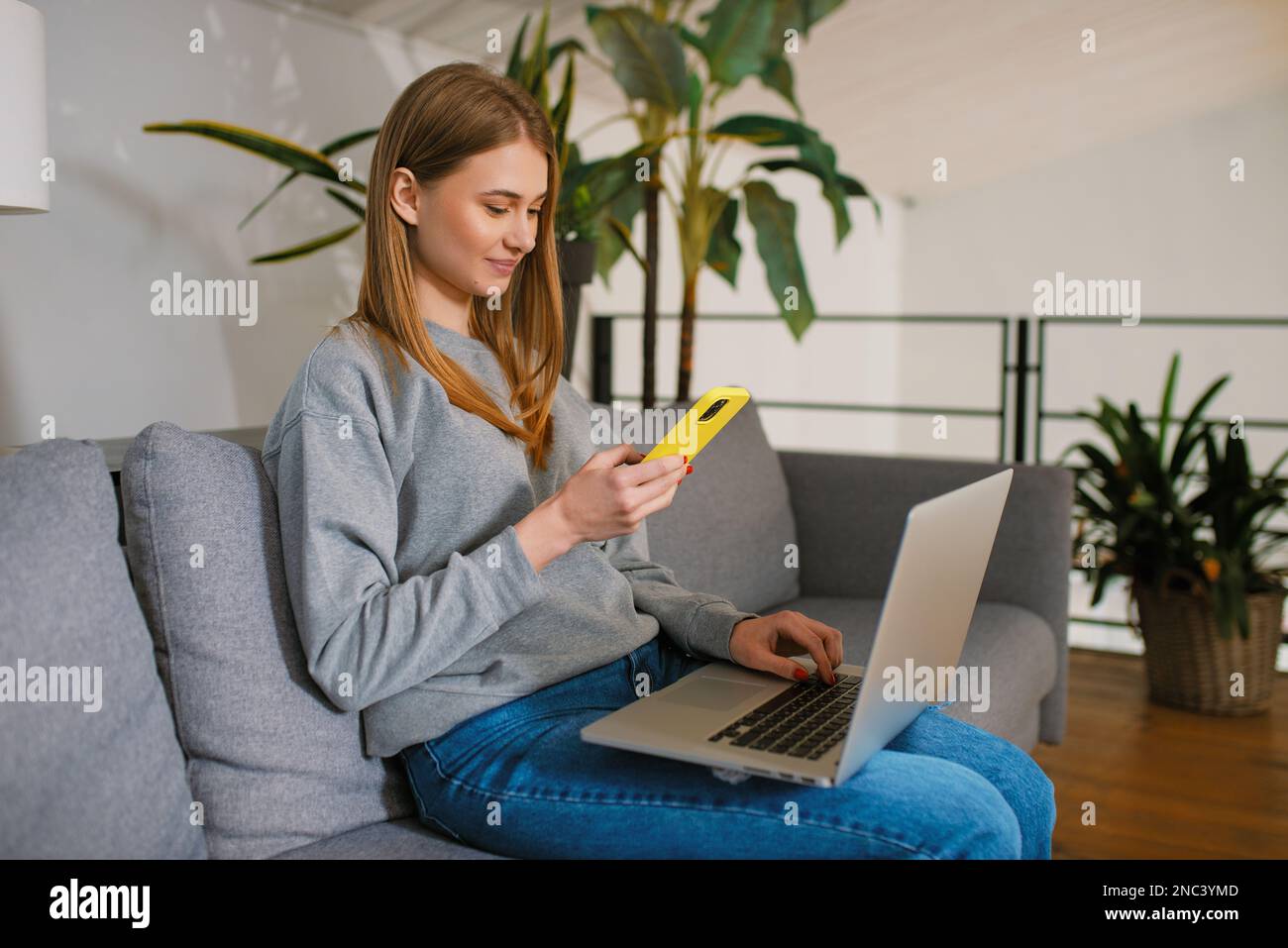 Casual happy young woman using smartphone in hand a looking a modern laptop screen sitting on cozy sofa at home, Hands at work with Digital Technology Stock Photo