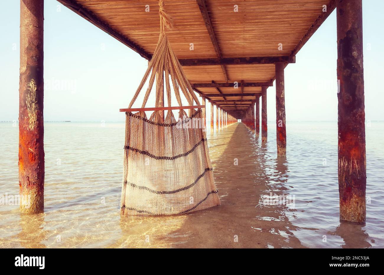 Hand woven natural fiber hammock under a pier, selective focus, color toning applied. Stock Photo
