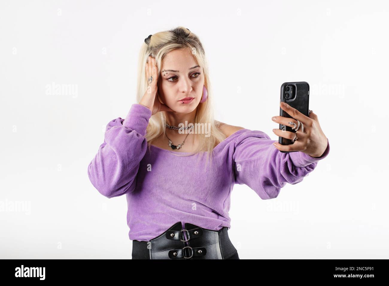 Young blonde woman wearing lilac ribbed blouse isolated over white background bored and holding head with a palm while video chatting with smartphone. Stock Photo