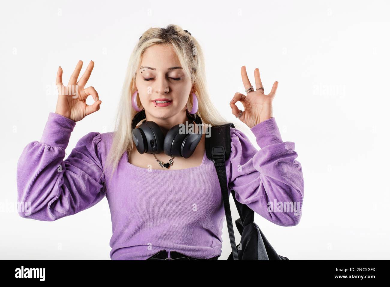 Portrait of young blonde woman wearing ribbed blouse standing isolated over white background with headphones around neck and eyes closed, doing okay g Stock Photo