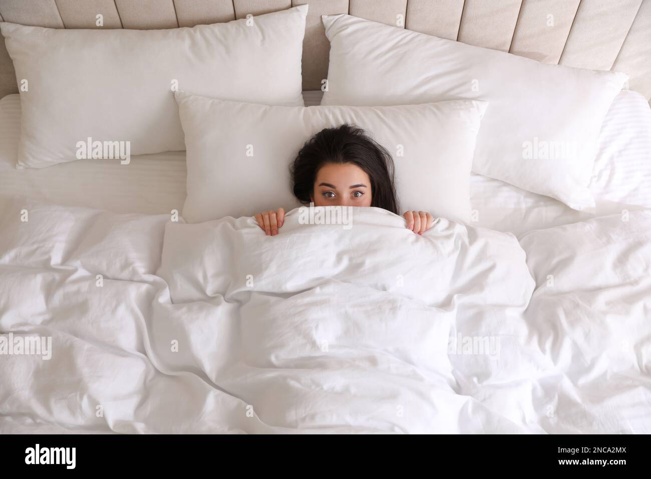 Young woman hiding under warm white blanket in bed, top view Stock