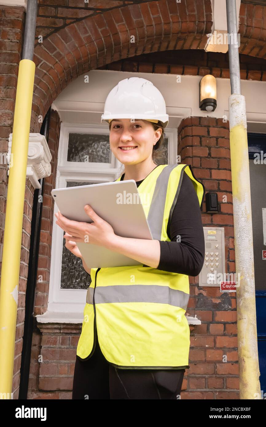 Vertical photo of a smiley female chartered civil engineer inspecting a construction site with a tablet, yellow high visibility vest, white hard hat Stock Photo