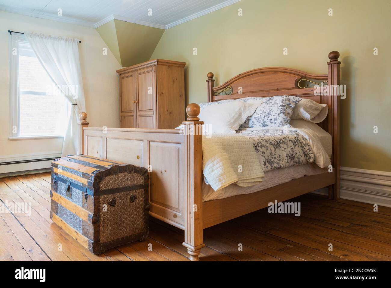 Queen size bed with wooden headboard and footboard, antique travel chest, armoire in upstairs master bedroom with oil stained fir wood floorboards. Stock Photo