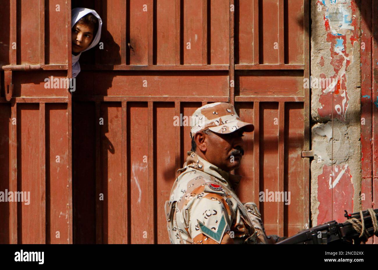 A Pakistani girl looks through a gate as a paramilitary soldier stands