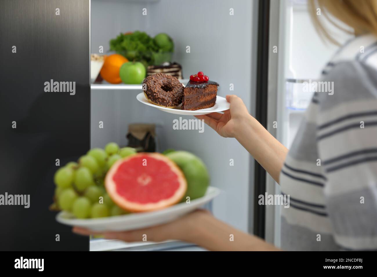 Choice concept. Woman holding plates with fruits and sweets near refrigerator in kitchen, closeup Stock Photo