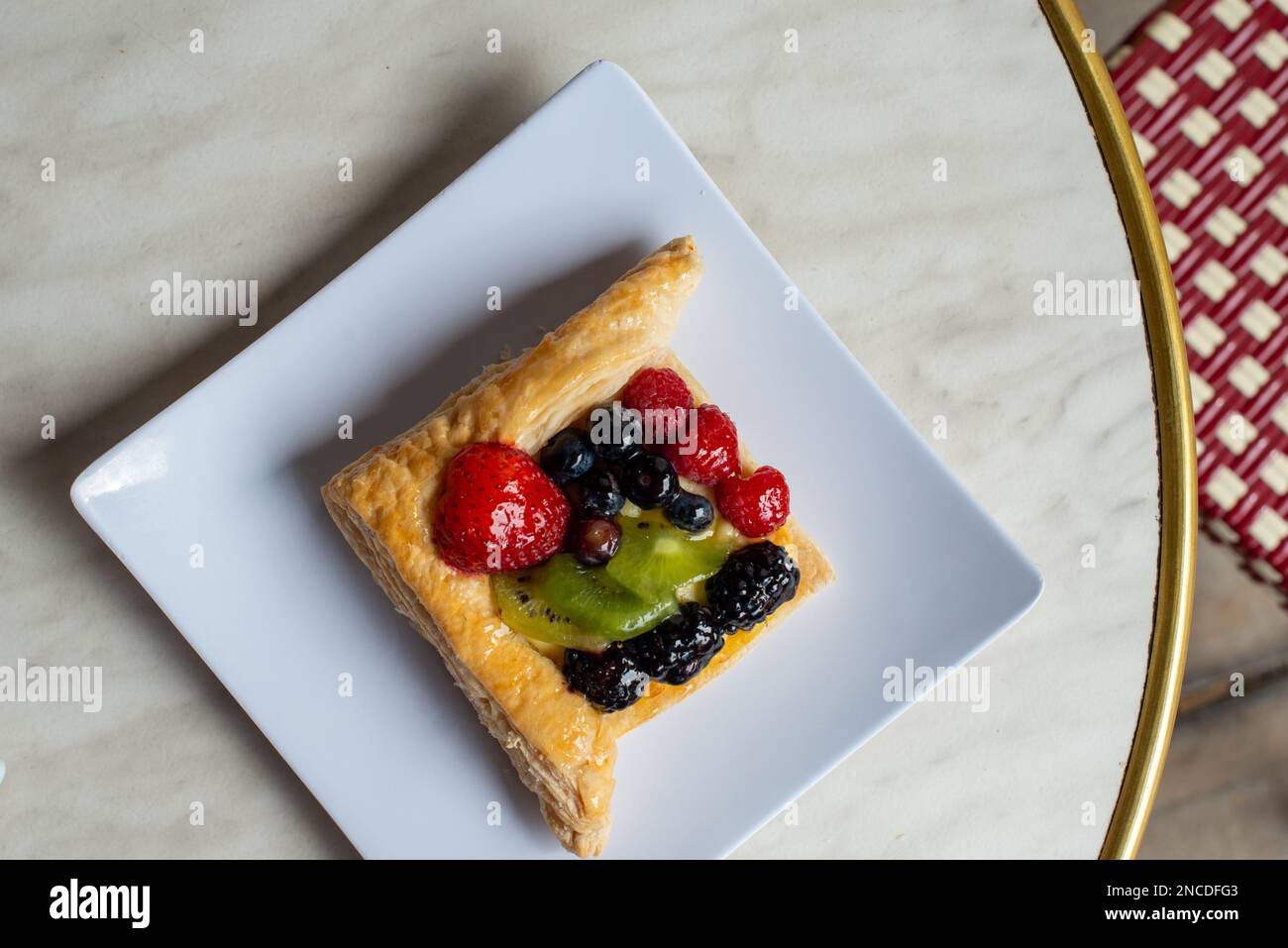 A square fruit filled French puff pastry on a white square plate. The cream and custard filled dessert has strawberries, kiwi, and blackberries on top Stock Photo