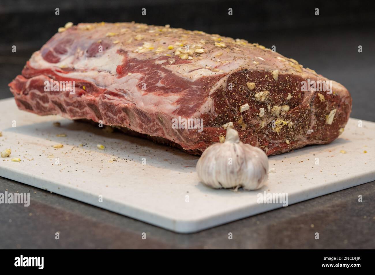 A large rack of raw beef prime rib on a white industrial plastic cutting board. The meat is sitting on the kitchen counter next to a  bulb of garlic. Stock Photo