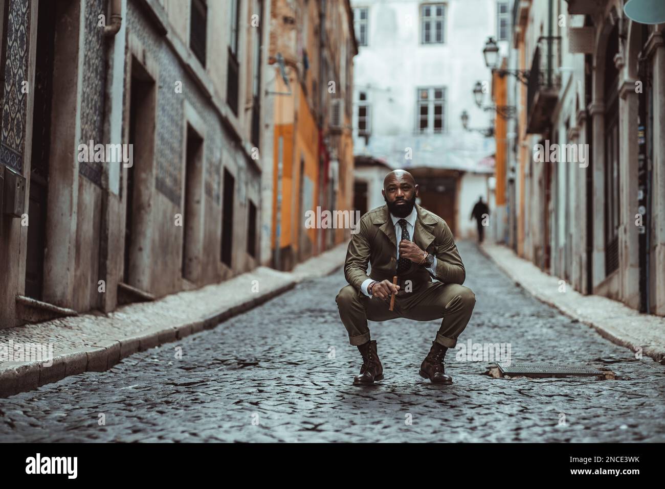 A shot focused on a bald black man dressed in khaki overalls standing in the middle of a black cobblestone street with typical old houses, crouching d Stock Photo
