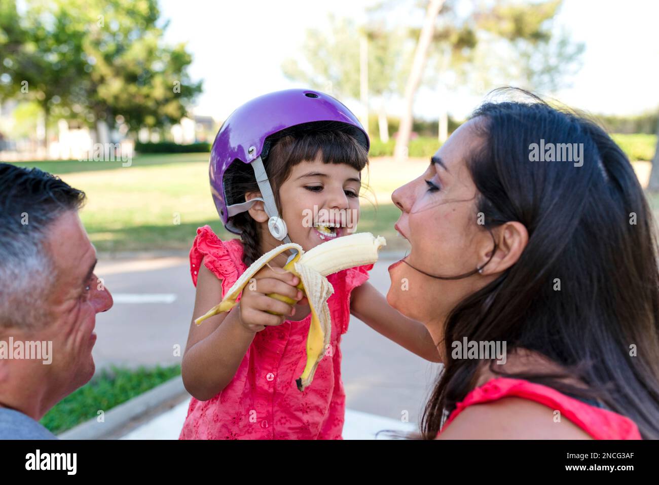 Little girl eating banana with parents sitting outdoors on the grass in a park. Stock Photo