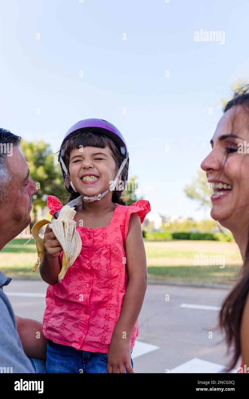 Little girl eating banana with parents sitting outdoors on the grass in a park. Stock Photo