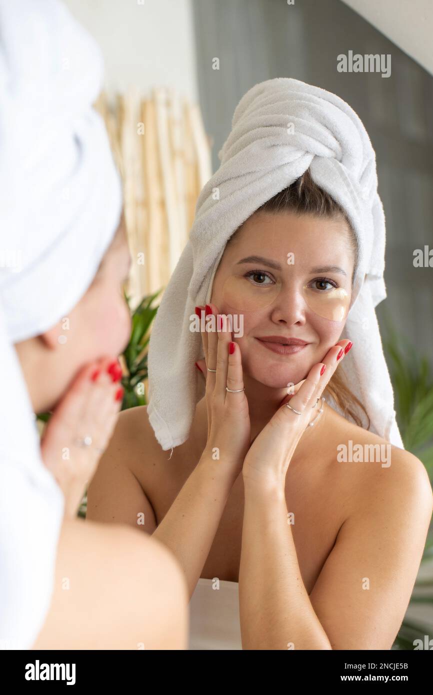 Attractive woman with patches under her eyes looks at her reflection in the mirror while smiling.  Stock Photo