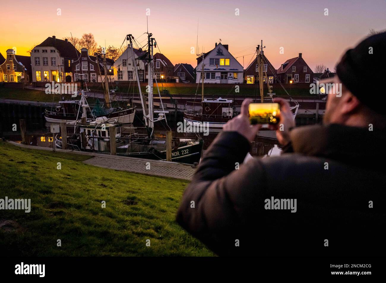 Greetsiel, Germany. 14th Feb, 2023. A tourist photographs shrimp cutters at sunset. The new North Sea Tourism Agency (Tano) wants to start building a uniform brand for the North Sea coast from the Ems to the Elbe this spring. (to dpa 'New tourism agency aims to strengthen sense of unity on the coast') Credit: Sina Schuldt/dpa/Alamy Live News Stock Photo