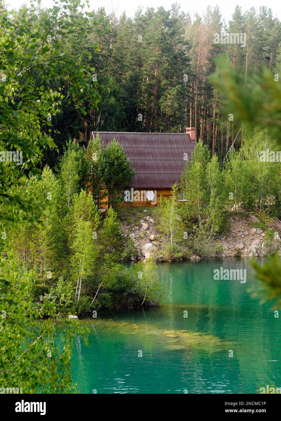 Wooden house with a satellite dish standing in the woods in the pine trees on the shores of stone lake with the island and turquoise water. Stock Photo