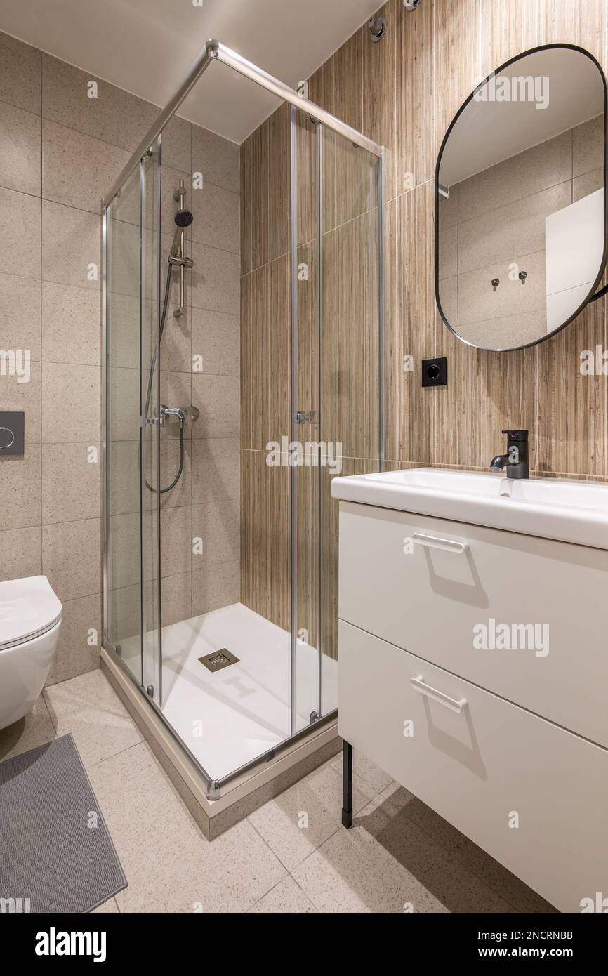 Bathroom with large shower area in corner. Rectangular washbasin on vanity table with white drawers and mirror on wall in black metal frame. Walls Stock Photo