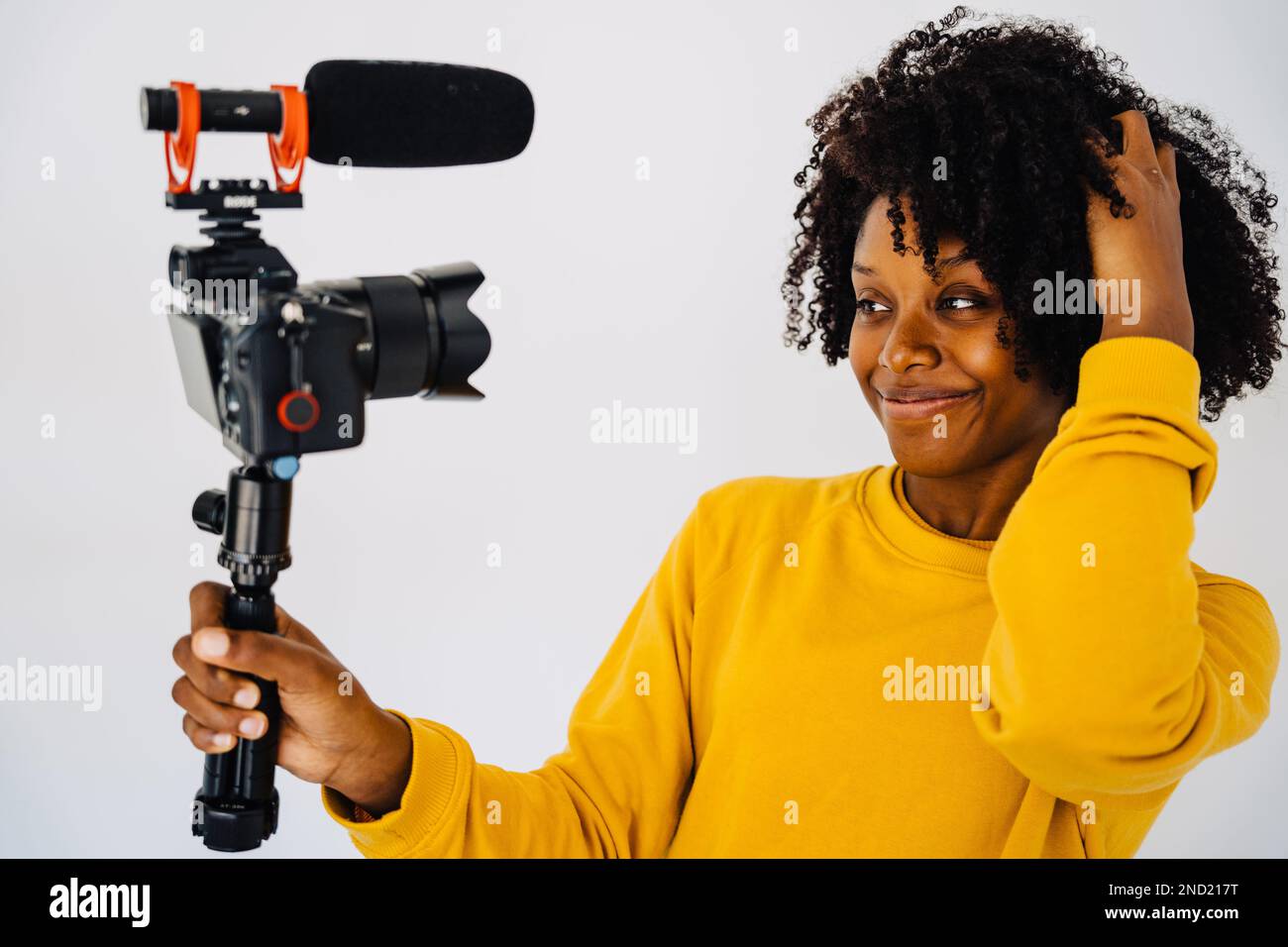 Confident smiling young African American curly haired female blogger in yellow sweatshirt standing against white wall and recording video on professio Stock Photo