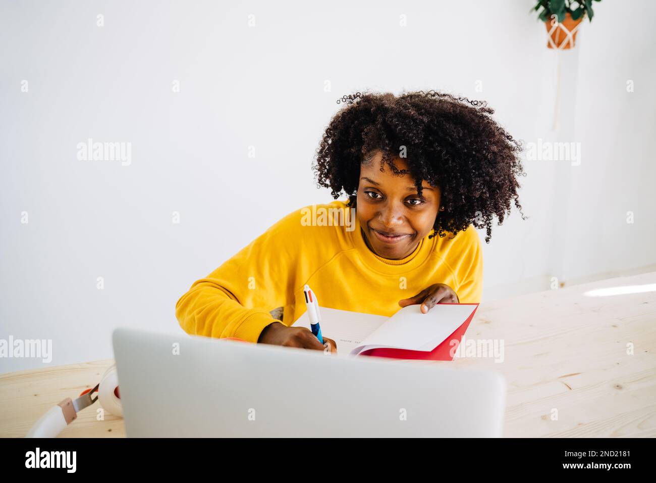 Positive young African American female student in casual outfit looking at screen of laptop while writing notes in copybook during exam preparation Stock Photo
