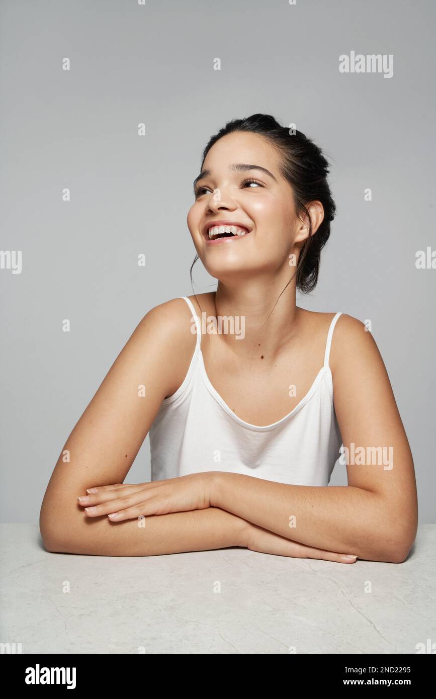 Positive young female in white top smiling and looking away while sitting against gray background with hands folded Stock Photo