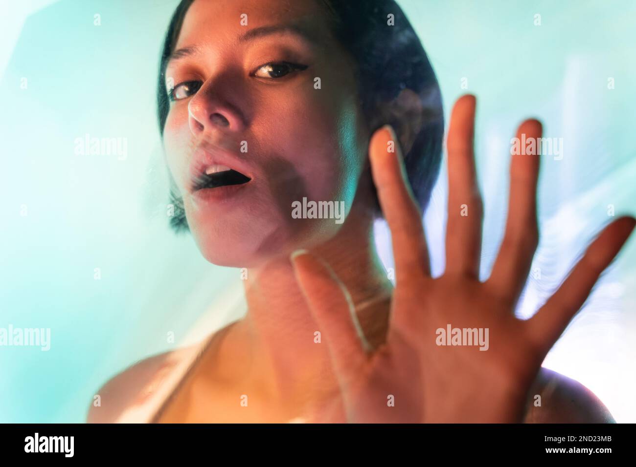 Through glass of confident young female with short hair and brown eyes looking at camera while showing stop gesture with mouth opened Stock Photo