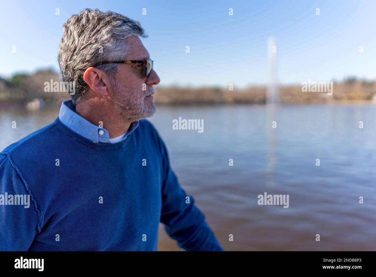Thoughtful elderly male in sunglasses and casual clothes standing on shore of lake while admiring picturesque scenery on sunny day Stock Photo