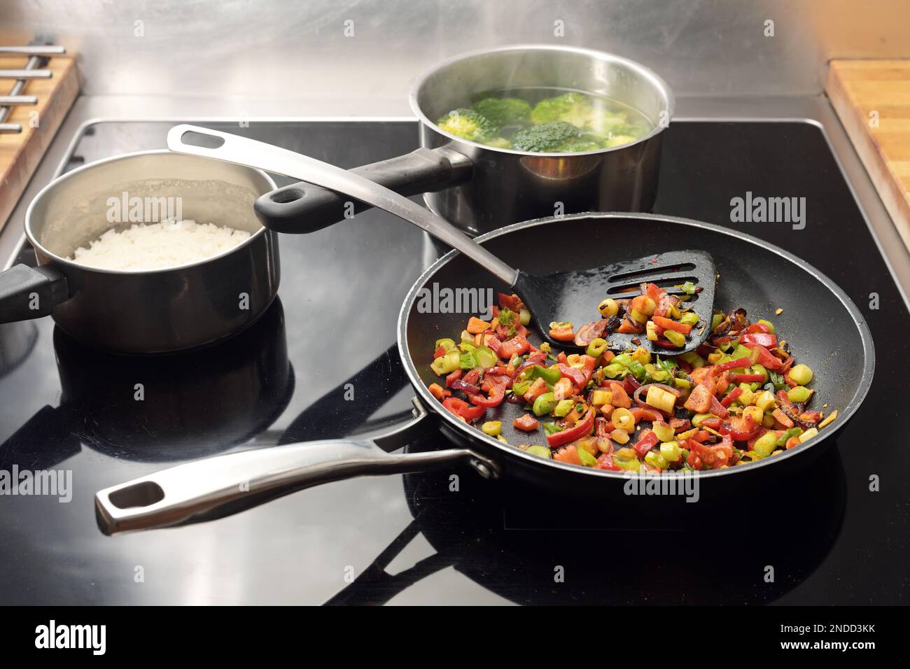 Diced vegetables are stir-fried with spices in a frying pan, pots with rice and broccoli behind them on the black stovetop, Asian style cooking, copy Stock Photo