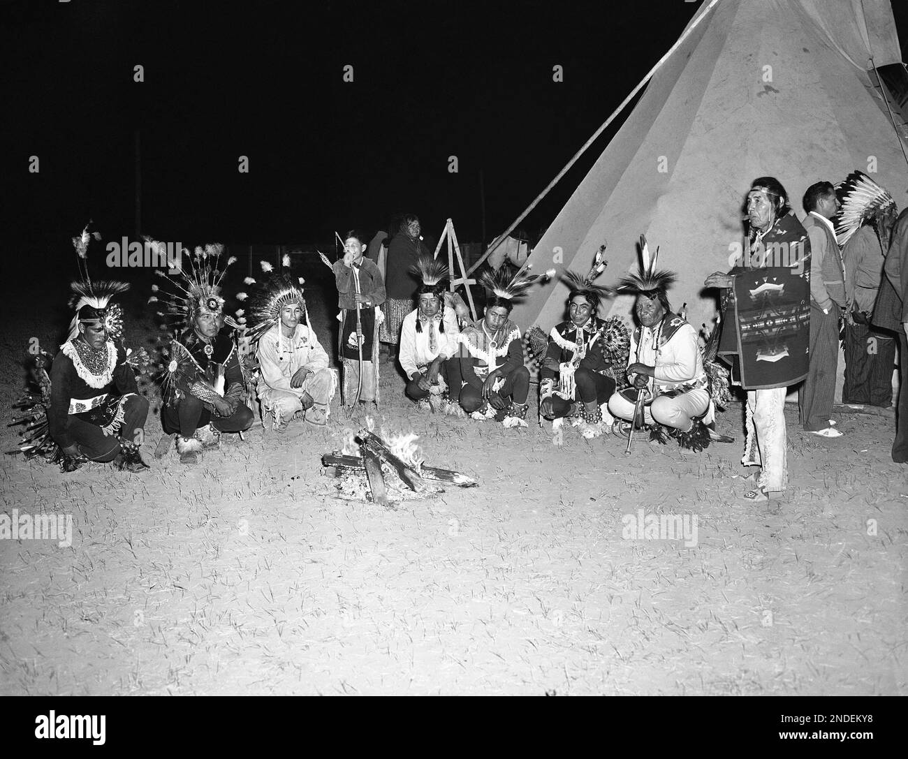 Dressed in full regalia, Shoshone Indians squat before a campfire ...