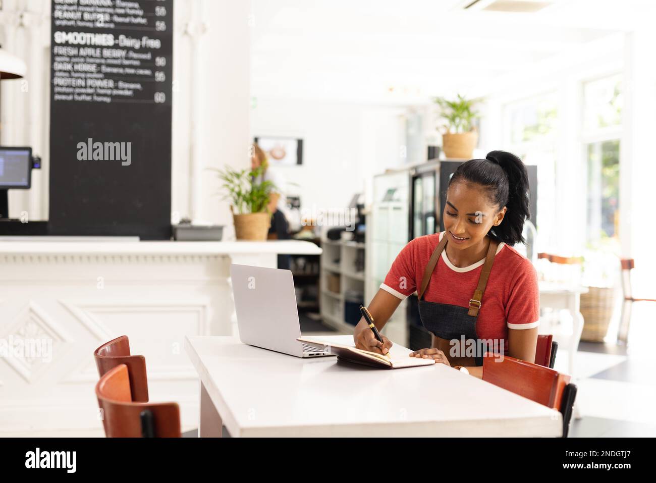 Happy biracial female barista wearing apron and taking notes in cafe ...