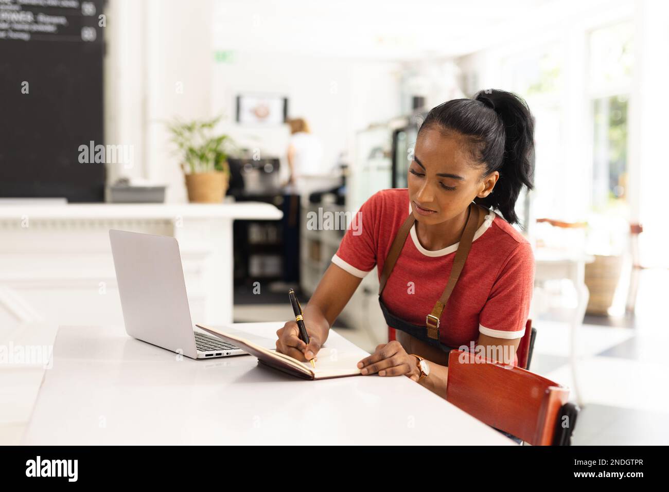 Happy biracial female barista wearing apron and taking notes in cafe ...