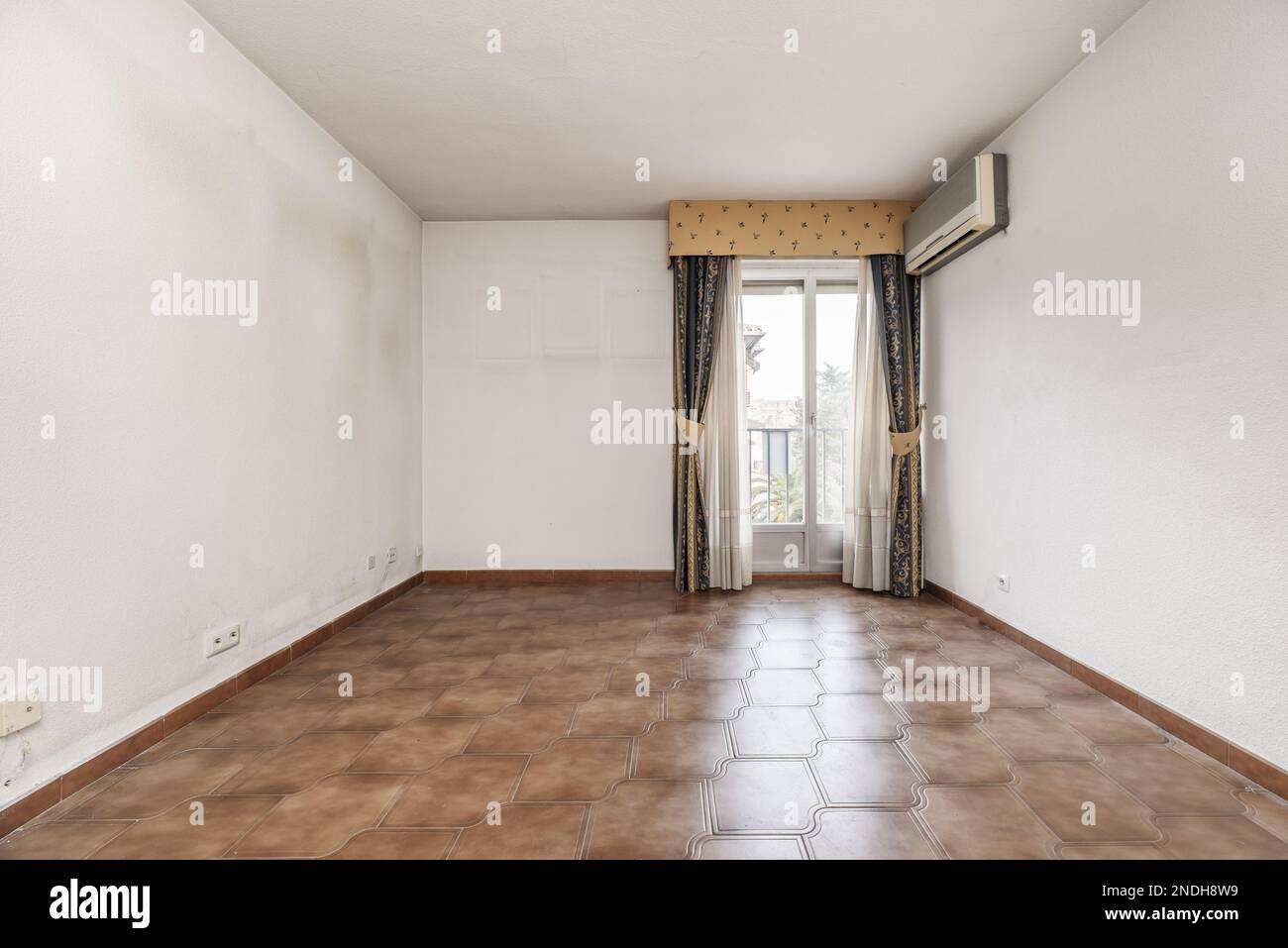 Empty room with old brown stoneware floor, wall mounted air conditioner and balcony with curtains and sheers Stock Photo