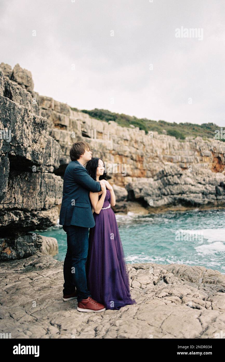 Man hugs woman from behind by the shoulders on the rocks by the sea ...