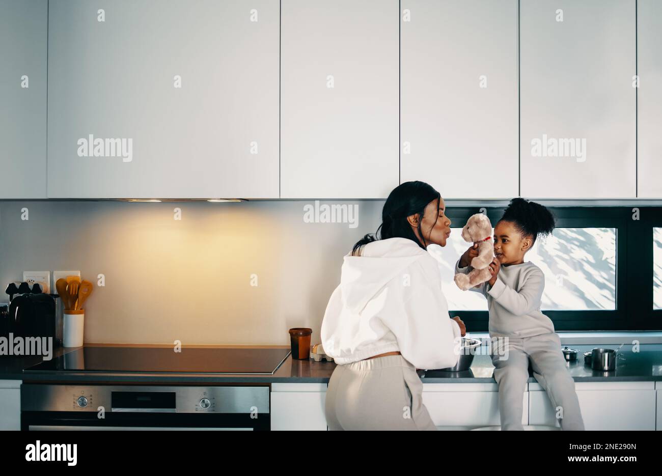 Mom playing with her daughter and her teddy bear in the kitchen. Woman giving a kiss to her child's toy as she is standing at the kitchen counter cook Stock Photo