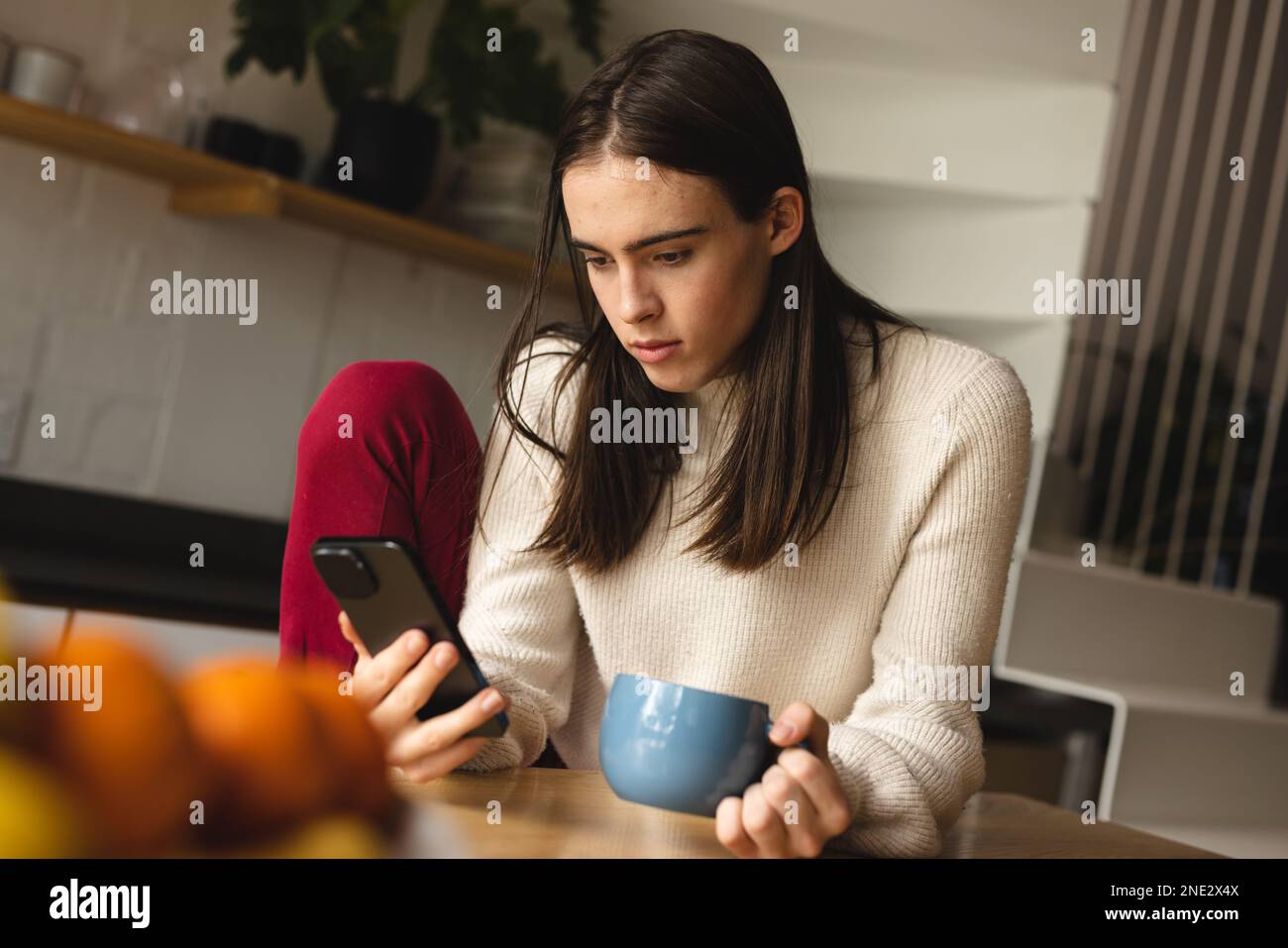 Non-binary trans woman holding a coffee cup using smartphone in the kitchen at home. concept of gender fluidity Stock Photo