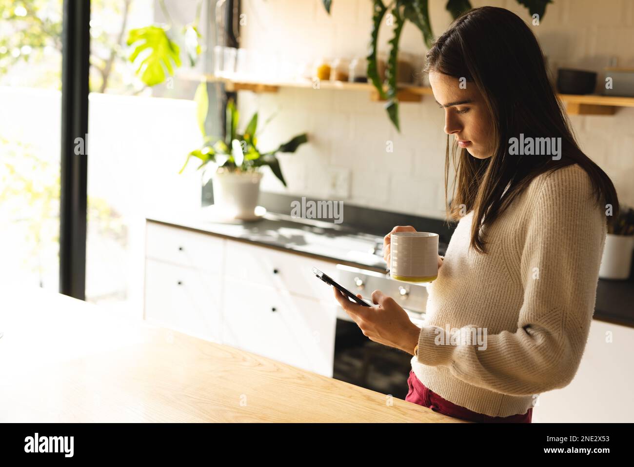 Non-binary trans woman holding a coffee cup using smartphone in the kitchen at home. concept of gender fluidity Stock Photo