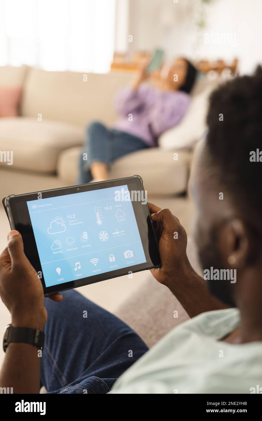 African american man sitting on sofa in living room and using tablet with smart home interface. Spending quality time at home, domestic life and lifes Stock Photo